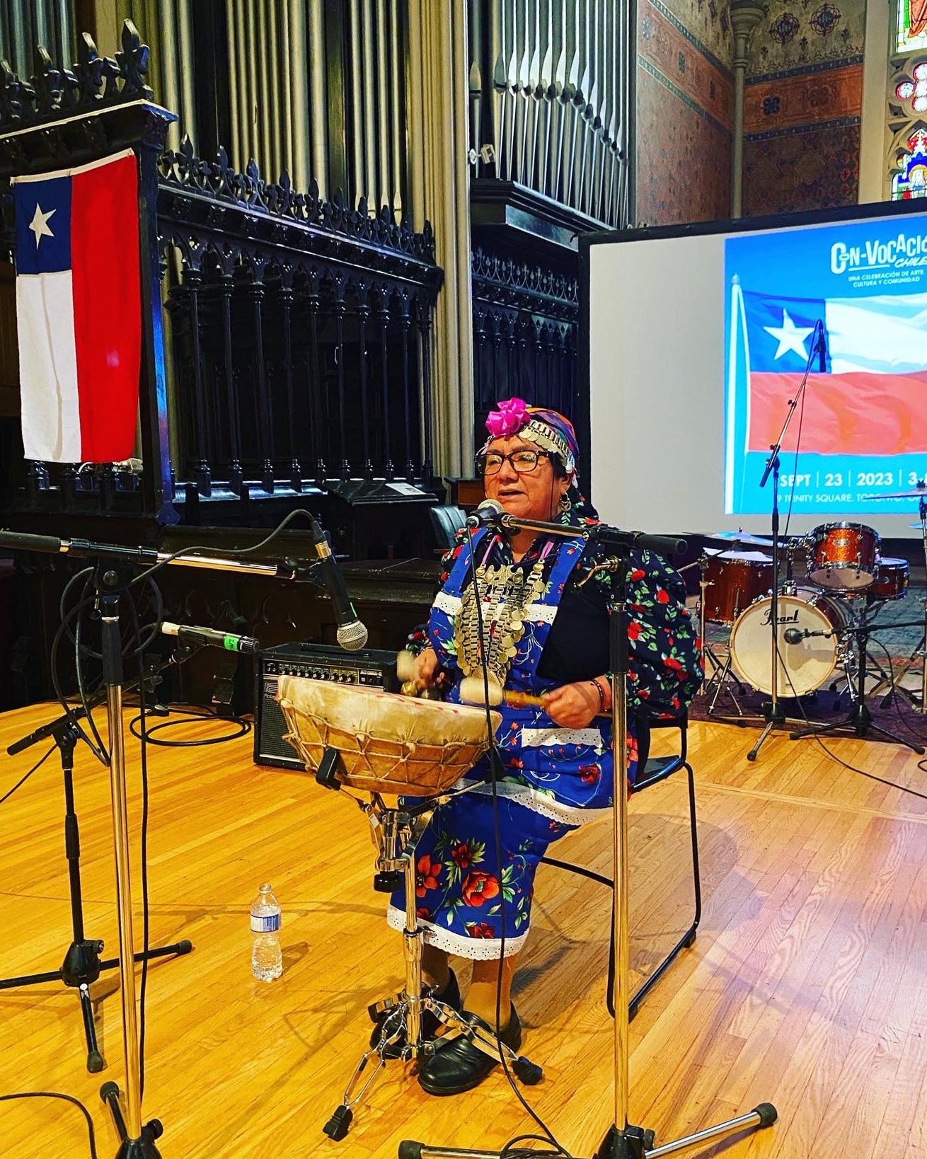 mujer mapuche cantando y tocando un tambor frente a unas banderas chilenas