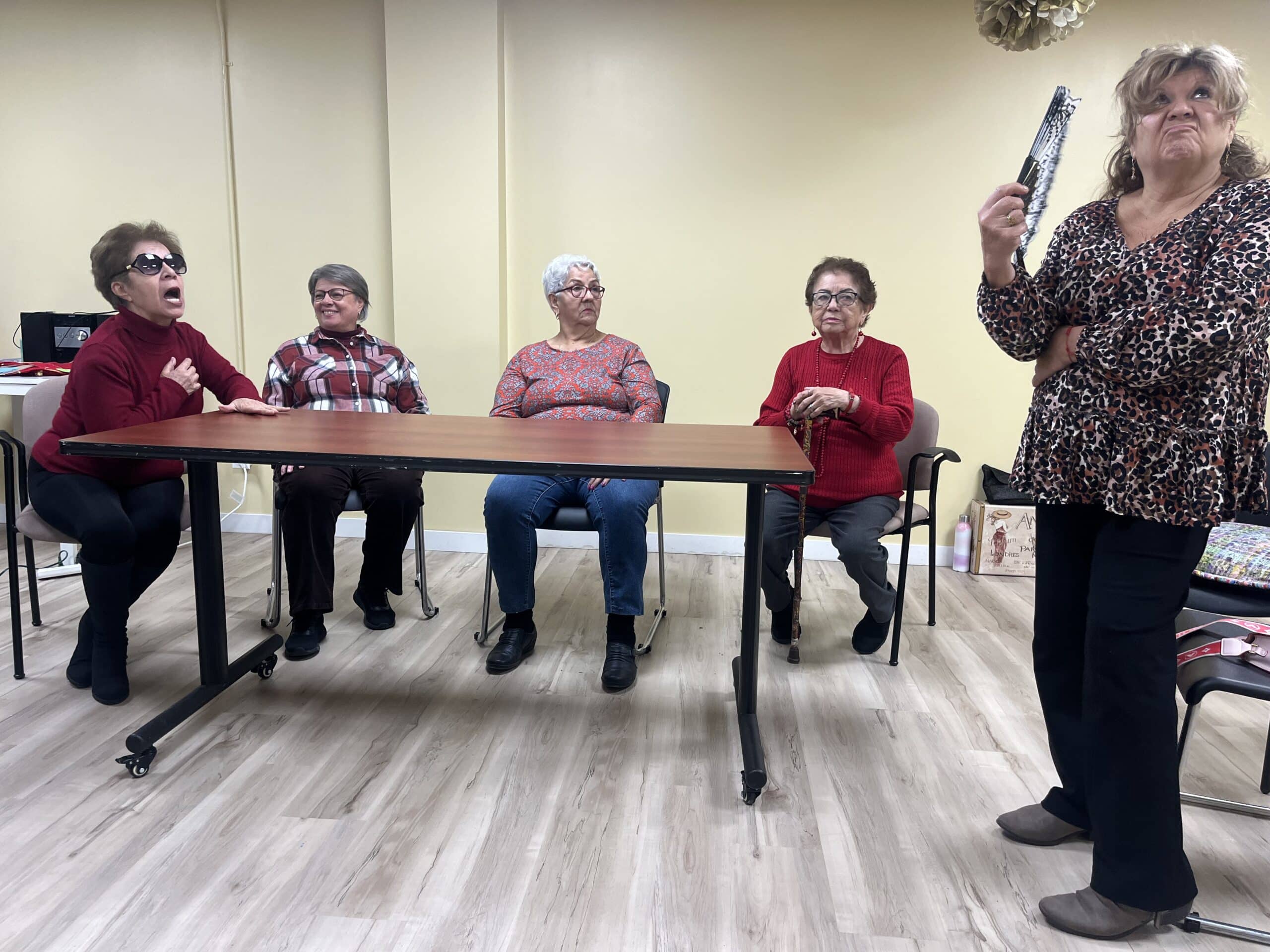 Cinco personas, cuatro de ellas sentadas frente a un escritorio y una de pie sosteniendo un abanico.